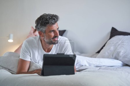 Photo for Mature Hispanic self employed man with curly hair using tablet and smiling while lying on bed and working remotely from home - Royalty Free Image