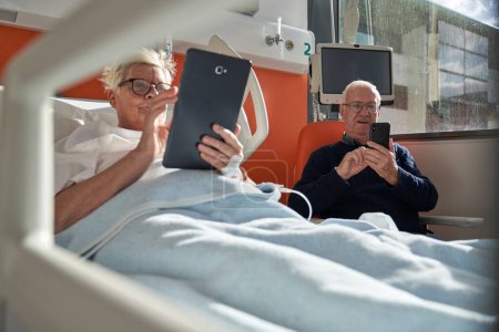 Foto de Hombre mayor serio sentado en sillón y mensajería en el teléfono inteligente cerca de la esposa enferma tumbado en el sofá y el uso de tabletas durante la rehabilitación en el hospital - Imagen libre de derechos