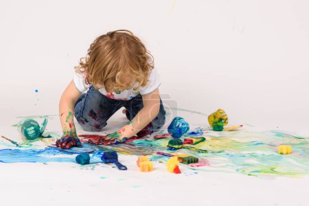Photo for Anonymous dirty boy smearing colorful paints on messy floor with empty plastic paint colors in light studio on white background - Royalty Free Image