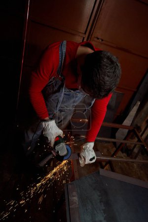 Photo for Young master in denim workwear leaning forward slightly while cutting metal railing by special tool during work - Royalty Free Image
