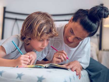 Téléchargez les photos : Adorable petite peinture de garçon avec des crayons colorés dans le carnet avec la mère tout en étant couché sur le lit - en image libre de droit