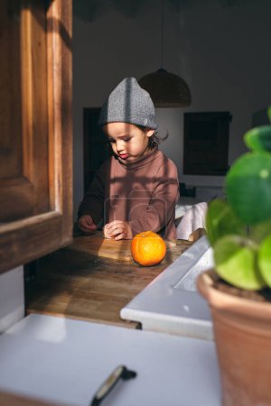Téléchargez les photos : Adorable petit garçon attentif dans des vêtements élégants debout à la table près de la fenêtre et peler des oranges dans la cuisine le jour ensoleillé - en image libre de droit