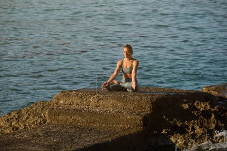Photo for Young female athlete taking Padmasana pose with hands in gyan mudra while sitting on boulder surrounded by rippling blue ocean and practicing yoga - Royalty Free Image