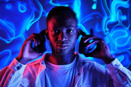 Téléchargez les photos : Homme afro-américain concentré portant un casque dans une pièce sombre avec un éclairage au néon vif et lumineux et regardant la caméra - en image libre de droit