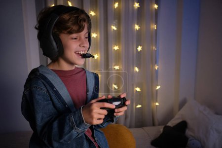 Photo for Side view of cheerful preteen child in headphones with microphone standing near bed with joystick and playing videogame in room decorated with glowing garland - Royalty Free Image