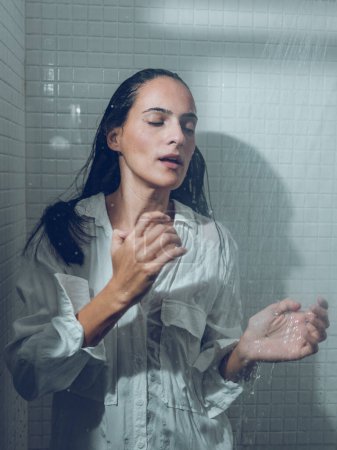 Téléchargez les photos : Attrayant hispanique femelle en chemise humide blanche debout avec les yeux fermés sous la douche éclaboussante dans la cabine dans la salle de bain - en image libre de droit