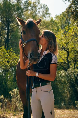 Foto de Contenido jinete femenino abrazar y besar caballo castaño en bozal mientras está de pie en el día soleado en el bosque en verano - Imagen libre de derechos