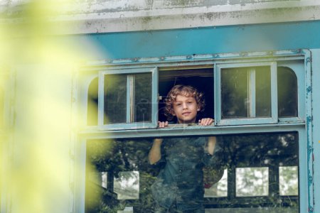 Photo for Delighted kid with curly blond hair looking out of opened window while standing in train and waiting for departure and looking away dreamily in sunny day - Royalty Free Image
