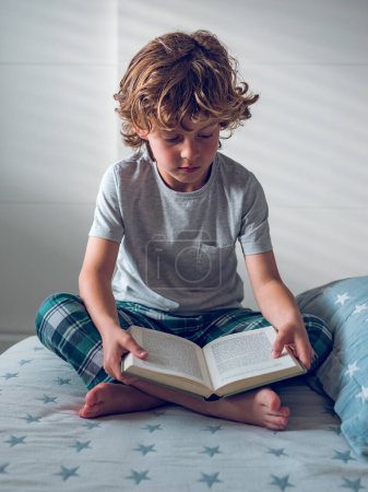 Photo for Full body of focused preteen boy in sleepwear sitting with crossed legs on soft bed and reading exciting story in book - Royalty Free Image