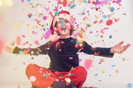 Photo for Full body of optimistic boy in Santa hat looking up with spread arms while sitting under colorful falling confetti during Christmas celebration - Royalty Free Image