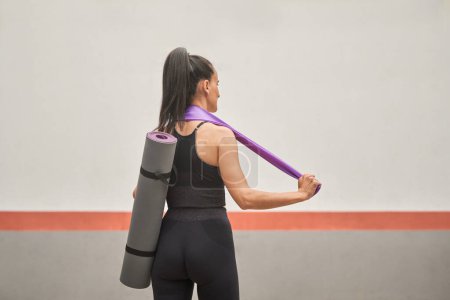 Téléchargez les photos : Vue de dos de femme en forme méconnaissable dans le soutien-gorge de sport et leggings debout en studio avec tapis de yoga roulé et bande élastique sur le cou après l'entraînement - en image libre de droit