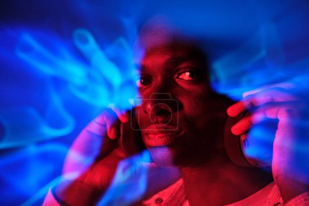 Foto de Hombre afroamericano en brillantes luces de neón escuchando música en auriculares y mirando a la cámara - Imagen libre de derechos