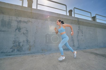 Téléchargez les photos : Jeune joggeuse en tenue de sport qui court vite et qui attend avec impatience sur la passerelle pendant l'entraînement le jour ensoleillé - en image libre de droit