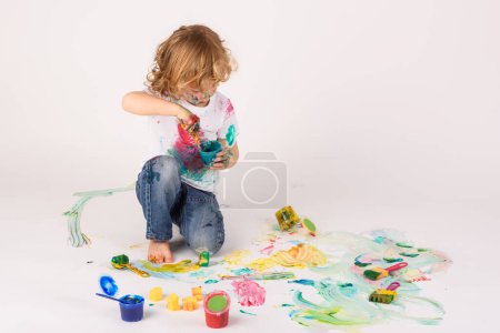 Foto de Cuerpo completo de chico sucio con lata de pintura sentada en el suelo desordenado mientras pinta en el estudio sobre fondo blanco - Imagen libre de derechos