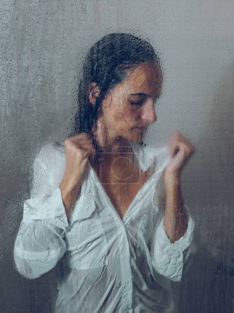 Foto de A través de vidrio de mujer infeliz en ropa blanca húmeda de pie en cabina de ducha con puerta transparente húmeda durante la rutina diaria - Imagen libre de derechos