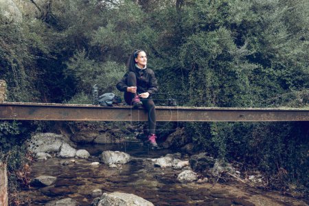 Foto de Cuerpo completo de joven y positiva excursionista hispana con cola de caballo en ropa deportiva descansando sobre un viejo puente metálico sobre un río rocoso y mirando hacia otro lado durante el trekking en el bosque de Mallorca - Imagen libre de derechos
