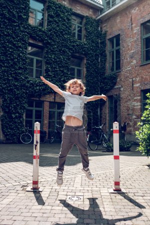 Photo for Full body of energetic kid with curly blond hair jumping with raised arms on path near shabby building with green plants on wall in sunny day - Royalty Free Image