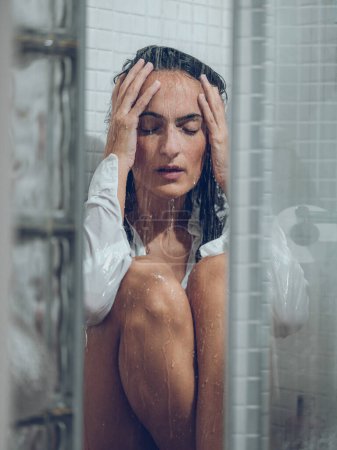 Photo for Lonely brunette with closed eyes in white wet shirt sitting in tiled shower under water and touching head gently - Royalty Free Image