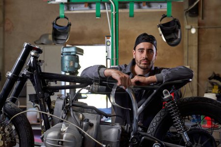 Téléchargez les photos : Technicien homme barbu confiant portant uniforme appuyé sur la moto en atelier et regardant la caméra - en image libre de droit