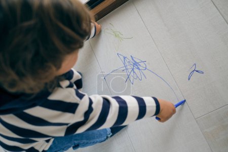 Téléchargez les photos : D'en haut d'anonyme chevelu bouclé bambin en vêtements décontractés dessin avec des marqueurs colorés sur sol stratifié près de la fenêtre - en image libre de droit
