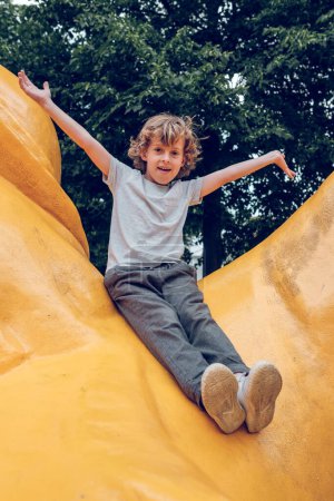Photo for From below full body of excited kid with curly hair and raised arms sitting on yellow stone sculpture in city park and looking at camera - Royalty Free Image