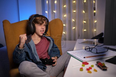 Photo for Preteen kid in headphones clenching fist while sitting in chair and celebrating success in video game in dim light - Royalty Free Image