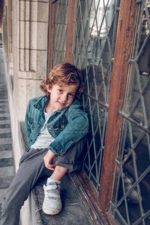 Téléchargez les photos : Enfant gai avec des cheveux bouclés dans la veste en denim assis sur le rebord de la fenêtre en pierre et posant sur la caméra - en image libre de droit