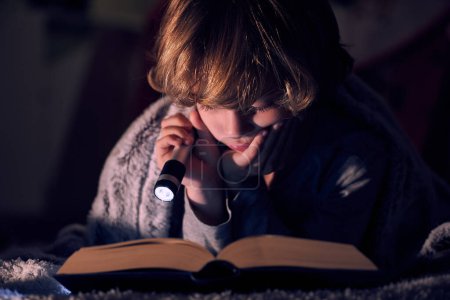 Foto de Libro de lectura niño tranquilo con linterna brillante mientras está acostado en la cama debajo de la manta en habitación oscura en la noche en casa - Imagen libre de derechos