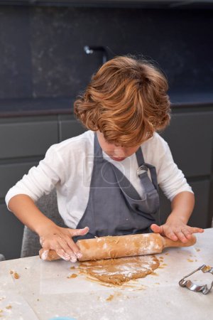 Téléchargez les photos : Petit garçon attentif aux cheveux blonds ondulés en vêtements décontractés et tablier pétrissant la pâte avec rouleau à pâtisserie en bois pendant la cuisson dans la cuisine - en image libre de droit