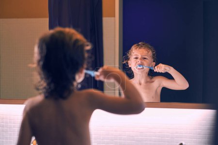 Téléchargez les photos : Vue arrière du garçon torse nu avec les cheveux mouillés réfléchissant dans le miroir et brossant les dents pendant la routine du matin dans la salle de bain - en image libre de droit