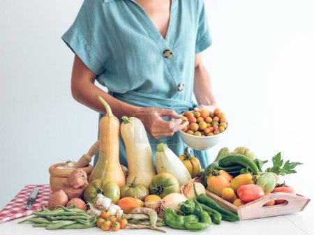 Téléchargez les photos : Cultiver une femelle anonyme debout près de la table avec des citrouilles fraîches et du poivre avec de l'oignon et de l'ail et tenant un bol de tomates sur fond blanc - en image libre de droit