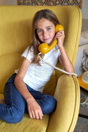 Photo for Smiling child sitting in armchair and holding handset of landline rotary telephone while looking at camera - Royalty Free Image