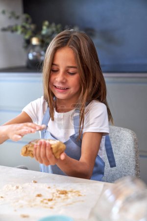 Photo for Glad girl in apron kneading raw cookie dough with hands while sitting at messy table in light kitchen of apartment - Royalty Free Image
