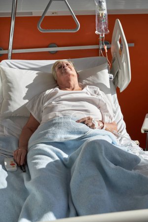 Foto de De arriba de enferma anciana paciente con pelo corto en ropa blanca acostada en cama médica con catéter venoso conectado y durmiendo durante el tratamiento en el hospital - Imagen libre de derechos