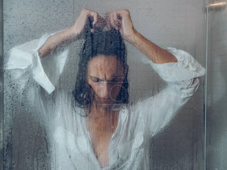 Foto de A través de vidrio hembra sin esperanza en ropa mojada tocando el cabello mientras está de pie cerca de la puerta transparente de la cabina de ducha en el baño ligero - Imagen libre de derechos