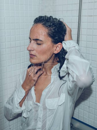 Photo for Thoughtful female in wet shirt standing with closed eyes under splashing water in shower cabin and touching head - Royalty Free Image