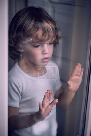 Photo for From above through glass of curious little boy with curly blond hair in white t shirt looking thought window at home - Royalty Free Image