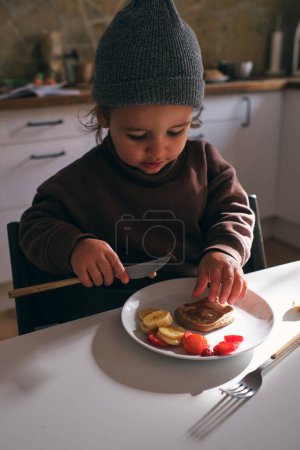 Photo for High angle of cute boy in hat sitting at table and eating bananas and strawberry with pancake - Royalty Free Image