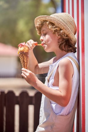 Téléchargez les photos : Vue latérale d'enfant aux cheveux bouclés en vêtements décontractés mangeant de délicieux cône de crème glacée avec bâton tout en s'appuyant sur un bâtiment coloré près de la clôture - en image libre de droit