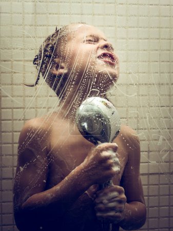 Téléchargez les photos : Petit enfant insouciant sans chemise avec les yeux fermés debout dans la salle de bain et tenant la pomme de douche avec de l'eau courante pendant la procédure d'hygiène - en image libre de droit