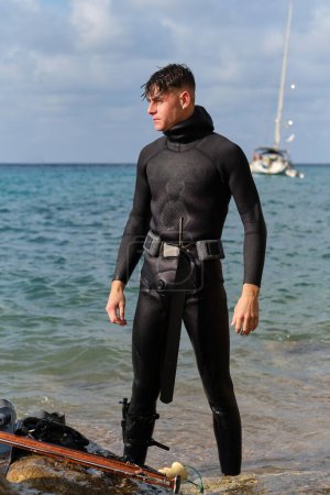 Photo for Determined male diver in wetsuit standing near gun on beach getting ready for spearfishing in sea and looking away against blurred sea - Royalty Free Image