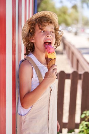 Téléchargez les photos : Vue latérale de l'enfant heureux dans l'ensemble et chapeau de paille léchant crème glacée rafraîchissante et regardant loin tout en se tenant près du mur rayé sur la rue de la ville le jour d'été - en image libre de droit