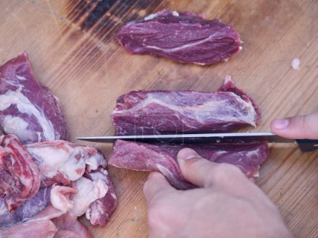 Téléchargez les photos : Vue du dessus du boucher mâle méconnaissable coupant du porc cru frais avec un couteau sur une planche de bois dans un abattoir - en image libre de droit