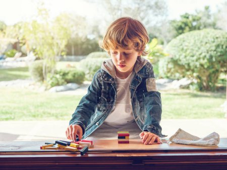 Téléchargez les photos : Adorable garçon concentré dans des vêtements décontractés jeu de tour tout en étant assis à la table en bois dans la cour arrière en été - en image libre de droit
