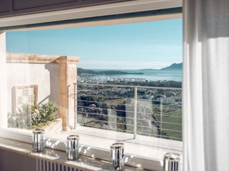 Téléchargez les photos : Par la fenêtre vue de la chambre d'hôtel donnant sur le balcon et le terrain vert et la mer azur par une journée ensoleillée à Pollensa ville de Majorque île - en image libre de droit