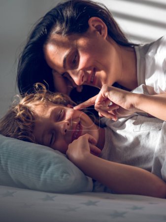 Téléchargez les photos : Joyeux toucher féminin joue de petit garçon souriant avec les yeux fermés tout en étant couché ensemble sur le lit à la maison - en image libre de droit