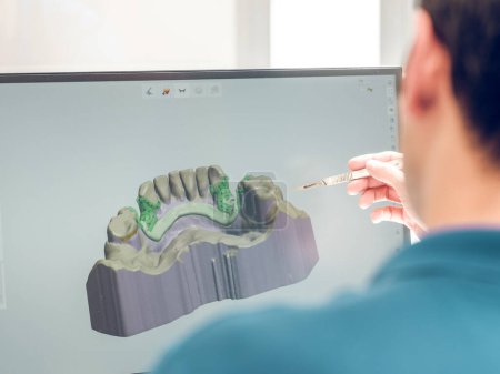 Téléchargez les photos : Vue arrière d'un stomatologue flou occupé et méconnaissable modelant la mâchoire sur ordinateur tout en travaillant dans une clinique contemporaine - en image libre de droit