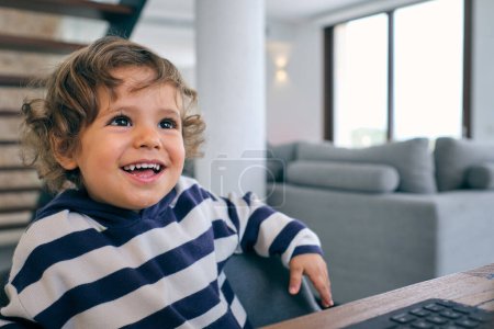 Téléchargez les photos : Mignon petit garçon assis à la table avec ordinateur portable et regardant loin tout en passant du temps dans le salon confortable à la maison - en image libre de droit