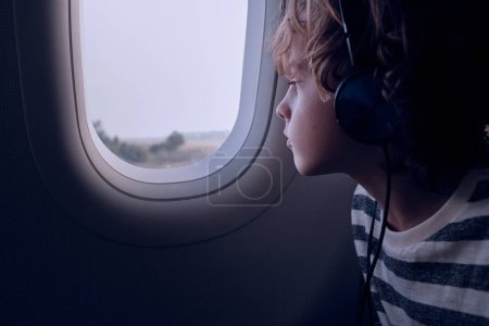 Photo for Kid listening to music in headphones and looking out window of dark plane during flight in evening - Royalty Free Image