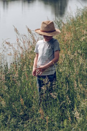 Téléchargez les photos : Enfant souriant méconnaissable en chapeau de paille debout parmi l'herbe verte haute près du canal calme et profitant de la journée d'été tout en regardant vers le bas - en image libre de droit